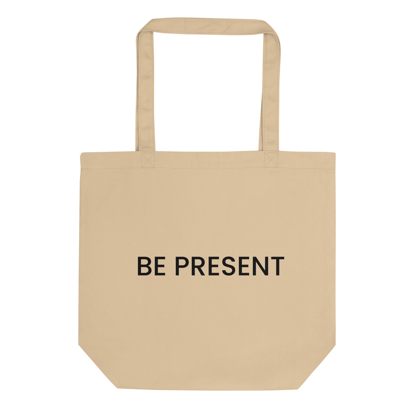 A light beige tote bag with the phrase "BE PRESENT" printed on the front in a bold, black, sans-serif font. The text is in all caps and centered on one line. The slogan serves as a powerful and simple reminder to practice mindfulness and focus one's attention on the current moment.