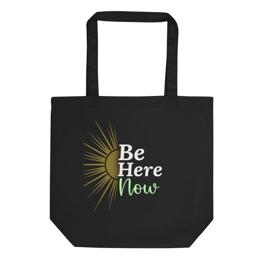 A black tote bag displays a stylized graphic of a rising sun on the left side. The sun is depicted with a semicircle in shades of gold and tan, emanating sharp, thin rays also in gold. To the right of the graphic, the empowering slogan "Be Here Now" is printed. "Be Here" is in a clean, bold white sans-serif font, stacked above "Now," which is written in a flowing, light green script font. This slogan encourages mindfulness and presence in the current moment.