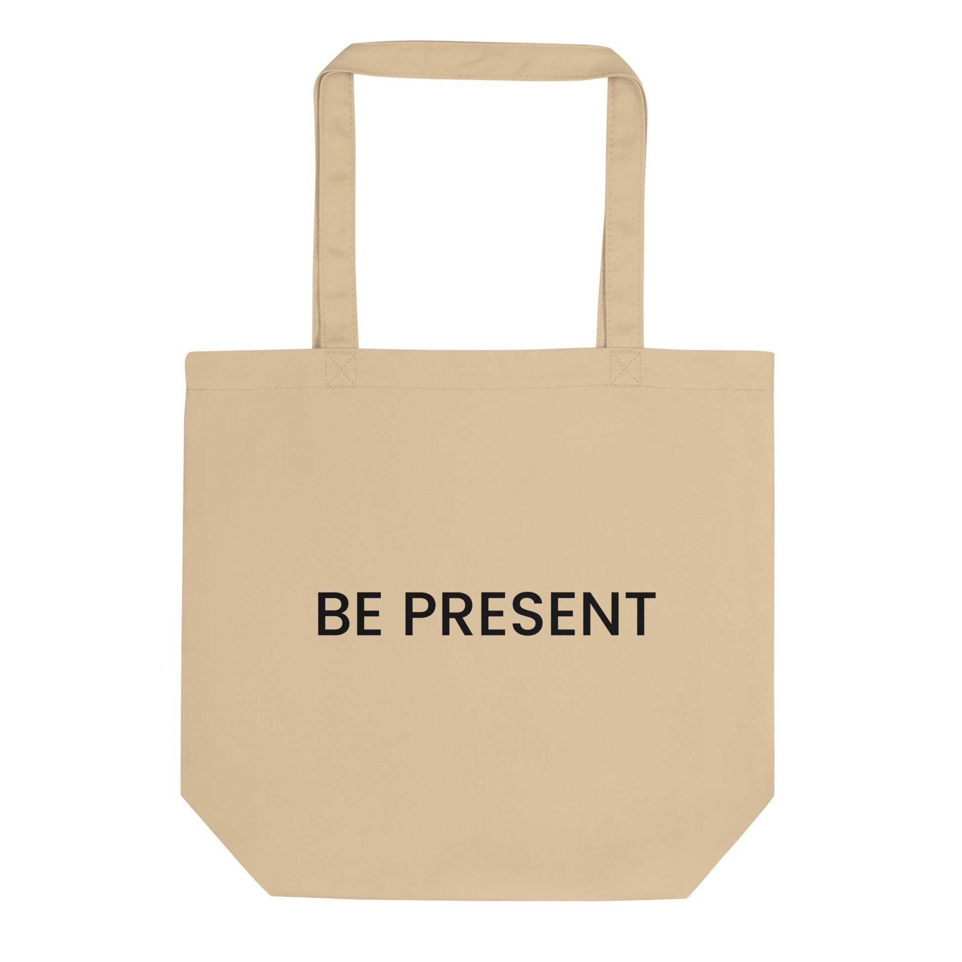 A light beige tote bag with the phrase "BE PRESENT" printed on the front in a bold, black, sans-serif font. The text is in all caps and centered on one line. The slogan serves as a powerful and simple reminder to practice mindfulness and focus one's attention on the current moment.