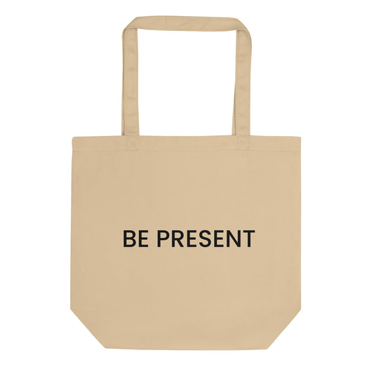 A light beige tote bag with the phrase "BE PRESENT" printed on the front in a bold, black, sans-serif font. The text is in all caps and centered on one line. The slogan serves as a powerful and simple reminder to practice mindfulness and focus one's attention on the current moment.