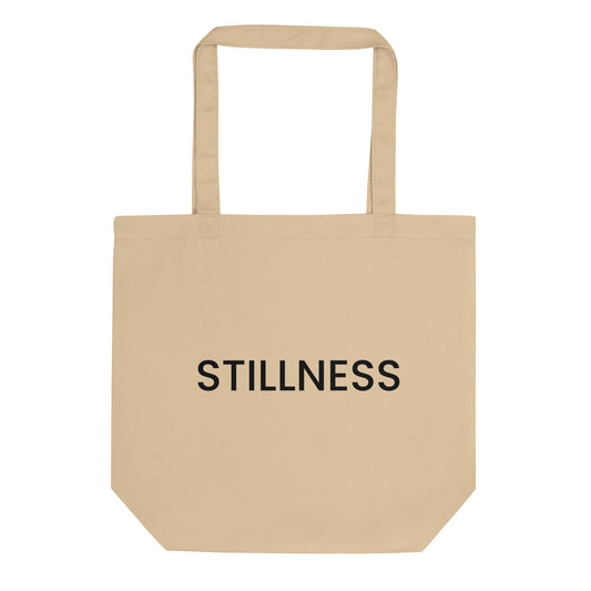A light beige tote bag with the word "STILLNESS" printed on the front in a bold, black, sans-serif font. The text is in all caps and centered on one line. The word represents a state of calm and quiet, encouraging a practice of finding peace amid the busyness of daily life.